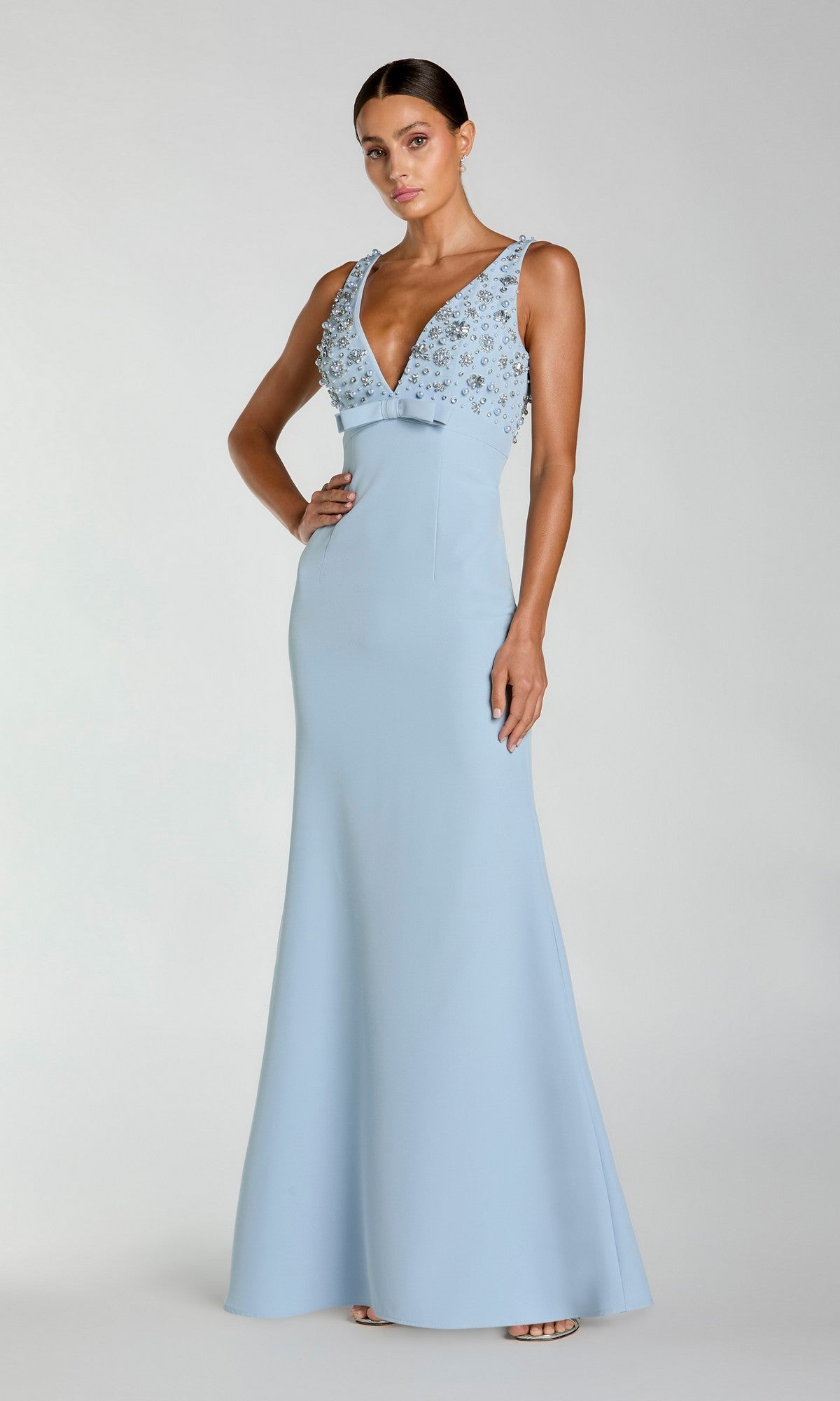 A woman models the Mac Duggal 2278 Beaded-Bodice Long Prom Dress, a sleeveless, floor-length light blue gown with a deep V-neckline and floral embellished beading, against a plain background.